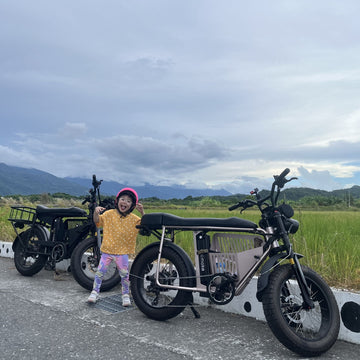 親子電輔車花東環島：小女孩戴粉紅色安全帽，站在Unimoke 胖胎電輔車中間，背景為壯麗山脈與綠意稻田