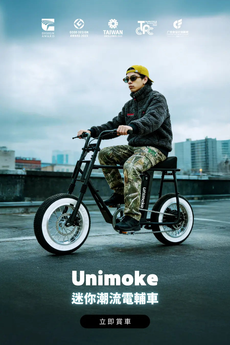 Unimoke 迷你潮流電輔車，黑色車身白色輪胎，一名男子穿迷彩褲、黑色抓毛外套、黃色帽子與墨鏡騎行於都市屋頂，呈現現代電動自行車潮流生活與設計感