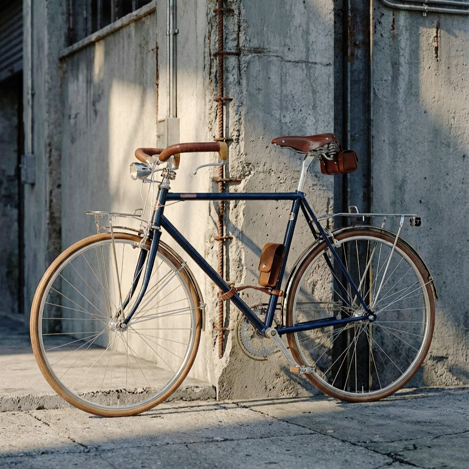 一台深藍色瑞士復古鋼管車，搭載車燈、彈簧式皮革座墊、皮革車架包、提把、貨架、泥除鏈蓋組與花梨木腳踏，在水泥牆前拍攝，復古質感一眼可見。