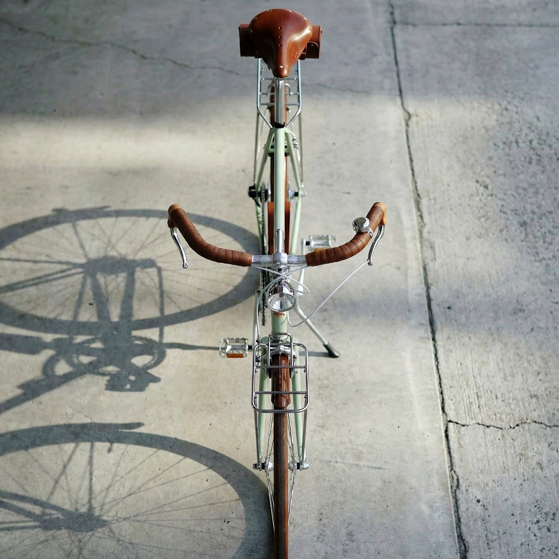 一台日系草芥綠瑞士復古鋼管車，俯視角度拍攝整車的樣貌，「全配備」設定，日常所需一次到位，成車升級包含 前燈、彈簧式皮革座墊、皮革車架包、提把、貨架、泥除鏈蓋組、尾燈、花梨木腳踏、真皮鎖包 ，置於灰色水泥牆前拍攝，質感低調卻很有存在感。